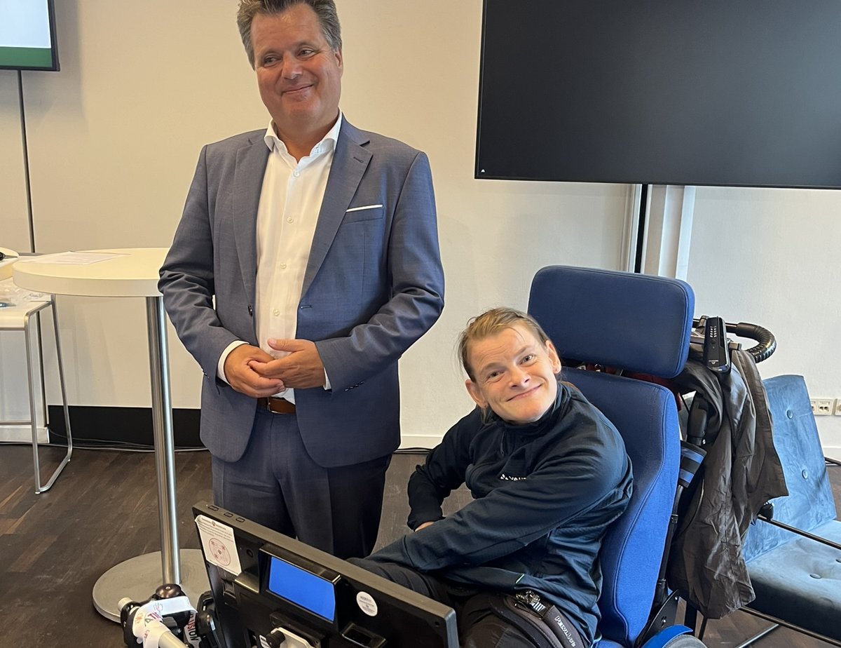 Nele Diercks im Rollstuhl an ihrem Sprachcomputer neben J&uuml;rgen Dusel, dem Beauftragten der Bundesregierung f&uuml;r die Belange von Menschen mit Behinderungen; beide l&auml;cheln in einem hellen Veranstaltungsraum