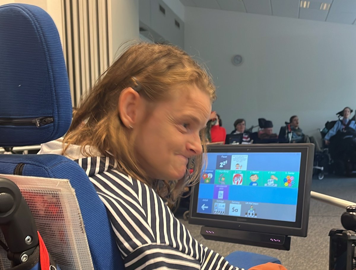 Nele Diercks im Rollstuhl an ihrem Sprachcomputer bei einem Treffen der UK-Referent:innen; im Hintergrund weitere Menschen im Rollstuhl