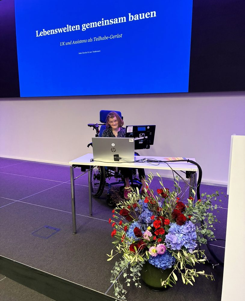 Ich auf der B&uuml;hne an meinem Sprachcomputer, hinter mir die Titelfolie &bdquo;Lebenswelten gemeinsam bauen&ldquo;