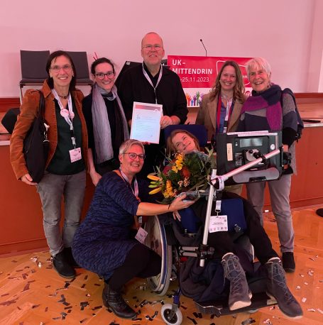 Nele Diercks mit Urkunde und Blumen bei der Preisverleihung, umringt von Wegbegleiterinnen, auf dem Kongress UK &ndash; mittendrin 2023
